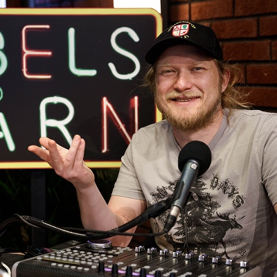 Person med caps og illustrert t-skjorte sitter foran en mikrofon i et lydstudio. Bakgrunnen har et neonskilt, og lydutstyr er synlig i forgrunnen.