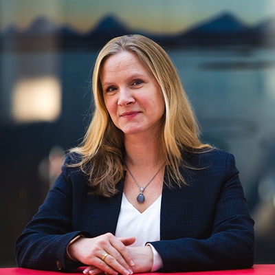 Bilde av Mari Sundli Tveit. Hun har langt hår og mørk blazer og sitter ved et bord i et kontormiljø.
