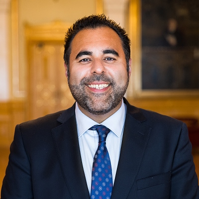 Masud Gharahkhani i mørk dress, hvit skjorte og mønstret slips. Han står innendørs foran en utsmykket tredør og vegg med innrammet kunst. Bildet er tatt i et formelt og elegant miljø