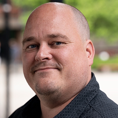 Bilde av Carl Henrik Knutsen. Han har skallet hode og har på seg en mørkfarget, mønstret skjorte. Bakgrunnen er utendørs med grønt løvverk og noen uskarpe strukturer.