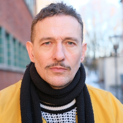 Bilde av Kristopher Schau. Han har på seg gul jakke, svart og hvit mønstret genser og svart skjerf og er fotografert i et urbant utendørsmiljø med bygninger og trær i bakgrunnen.