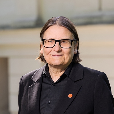 Person med briller og mørkt skulderlangt hår, kledd i svart skjorte og svart dressjakke, står foran en lys murvegg.