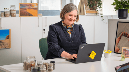 En smilende dame sitter i et kontorlandskap foran en PC og ser ned på skjermen