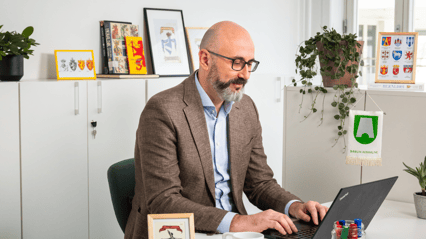 En mann med skjegg sitter i et kontorlandskap foran en PC.