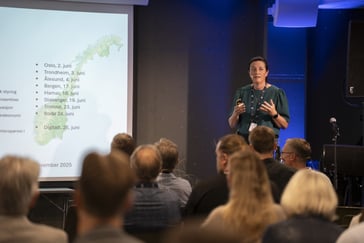en kvinne i grønn bluse står på en talerstol foran en presentasjon