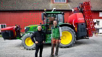 to menn i arbeidsantrekk står foran en grønn traktor og en rød låve