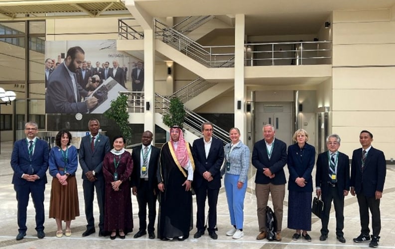 Gruppebilde av medlemmer fra styret i Global Research Council, stående på rekke innendørs foran en trapp og et stort veggbilde. Personene har navneskilt og bærer formelle klær, inkludert nasjonale drakter fra ulike land. Bildet viser mangfold og internasjonalt samarbeid.