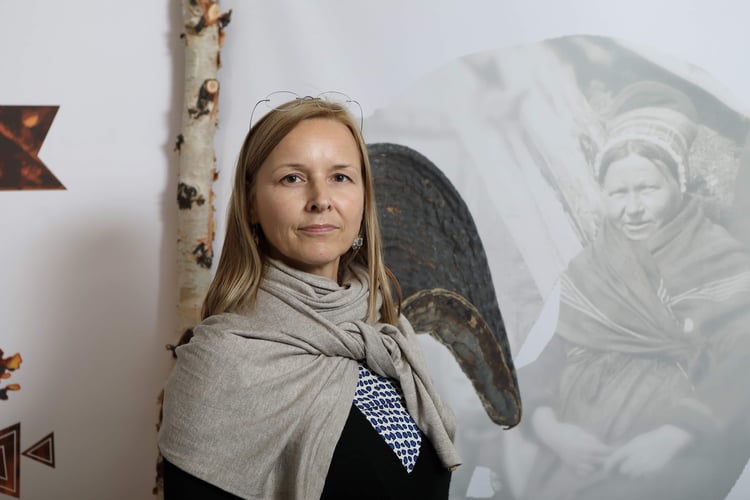 en kvinne med lyst hår, briller på hodet har stilt seg opp foran en utstilling i et museum, et bilde av en samisk kvinne i bakgrunnen og en bjørkestamme