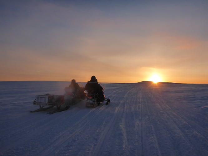 Two researchers on snowmobiles driving into a sunset