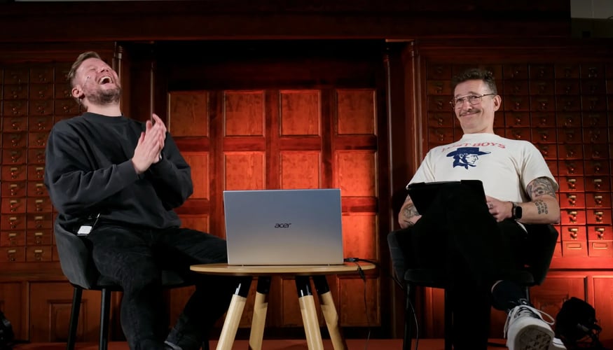 To menn sitter i hver sin stol på en scene med mørk bakgrunn. Mellom seg har de et bord med en bærbar PC. En lener ser bakover mens han ler, mens den andre ser lurt i kamera med et smil. 