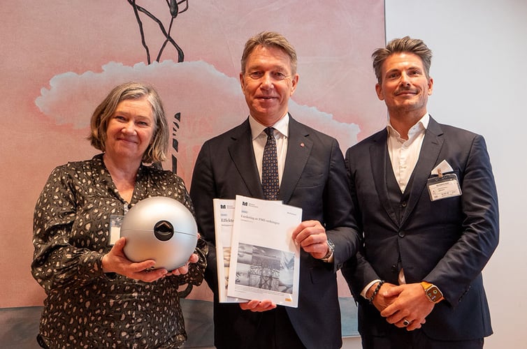 Tre personer poserer foran et maleri. Personen til venstre holder en rund, sølvfarget gjenstand, mens personen i midten holder to rapporter merket med logoen til Norges forskningsråd. Alle tre er pent kledd og smiler mot kameraet, trolig i forbindelse med en offisiell overrekkelse eller presentasjon.